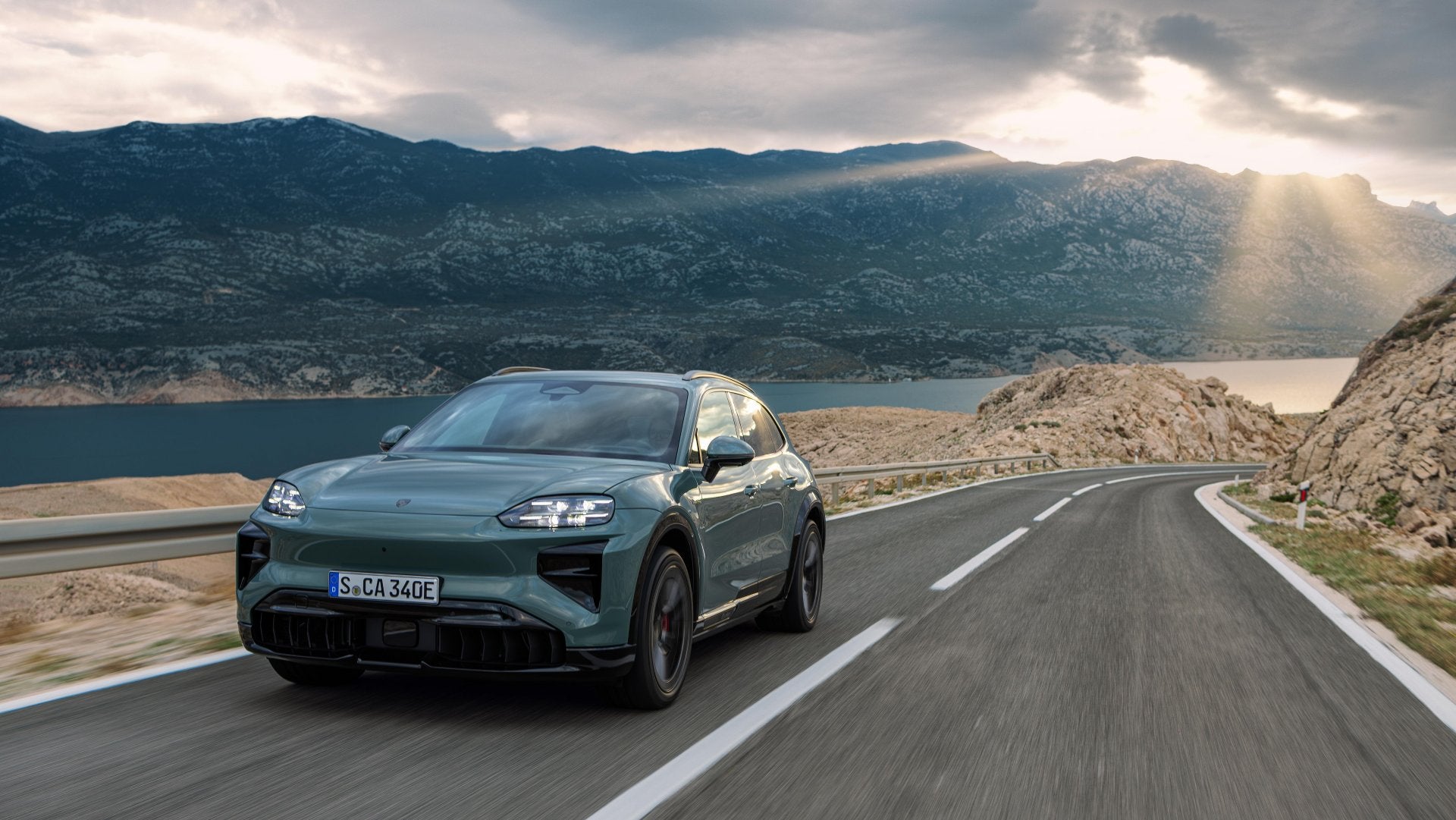 2026 Porsche Cayenne Turbo Electric driving through a twisting mountain road with dramatic sunlight breaking through the clouds.